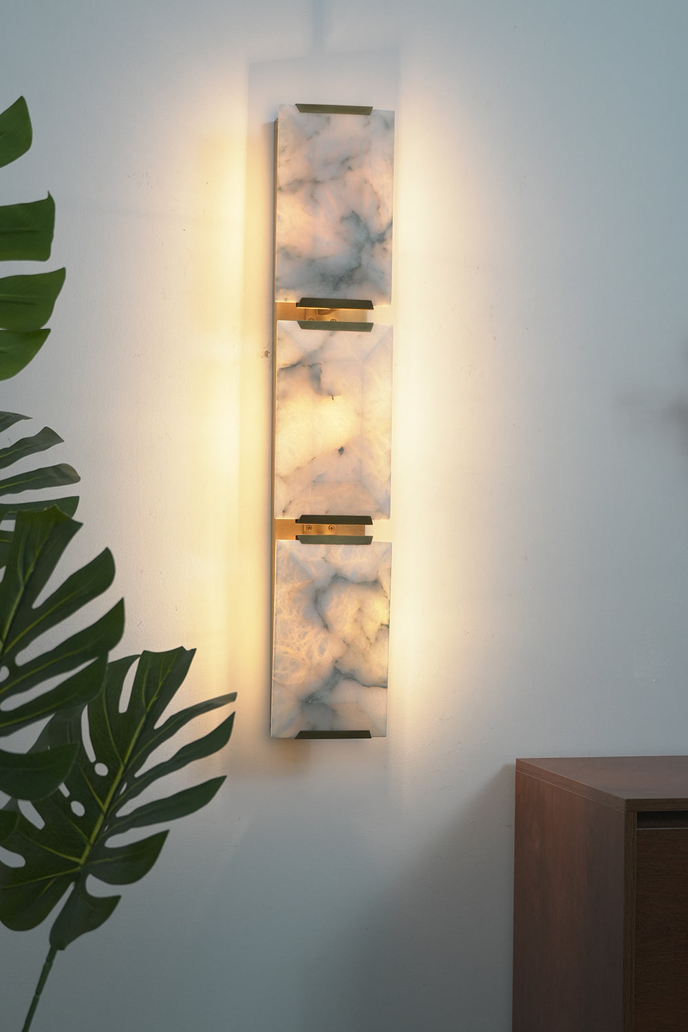 Lit 3-panel alabaster wall lamp | Brass, Alabaster | Perspective view against a white wall with indoor plants | Samu Lamp