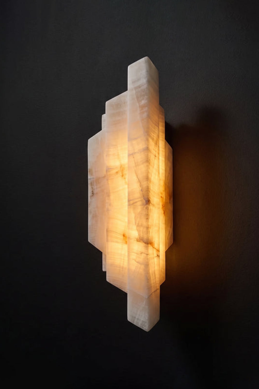 Alaric Tiered Alabaster Wall Sconce | Natural Alabaster and Metal | Angled side view of the illuminated multi-tiered geometric stone fixture against a dark black background | Samu Lamp