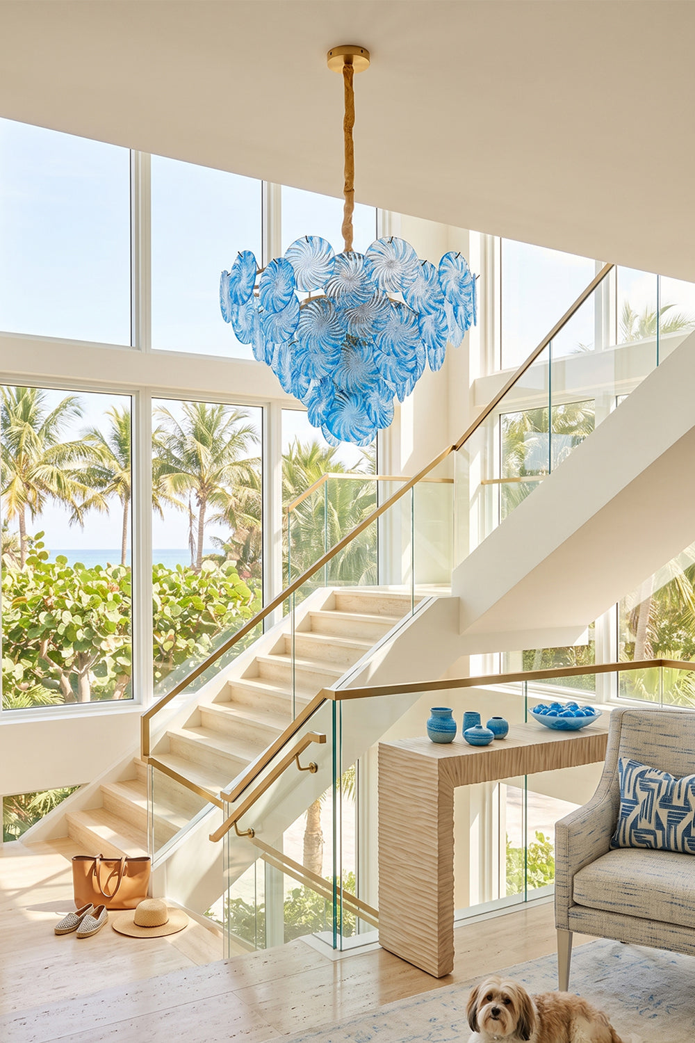 Amara Pink Glass Petal Chandelier | Staircase Foyer Light | Blue glass variant suspended as a grand statement ceiling light near a modern staircase with a scenic beach backdrop | Samu Lamp