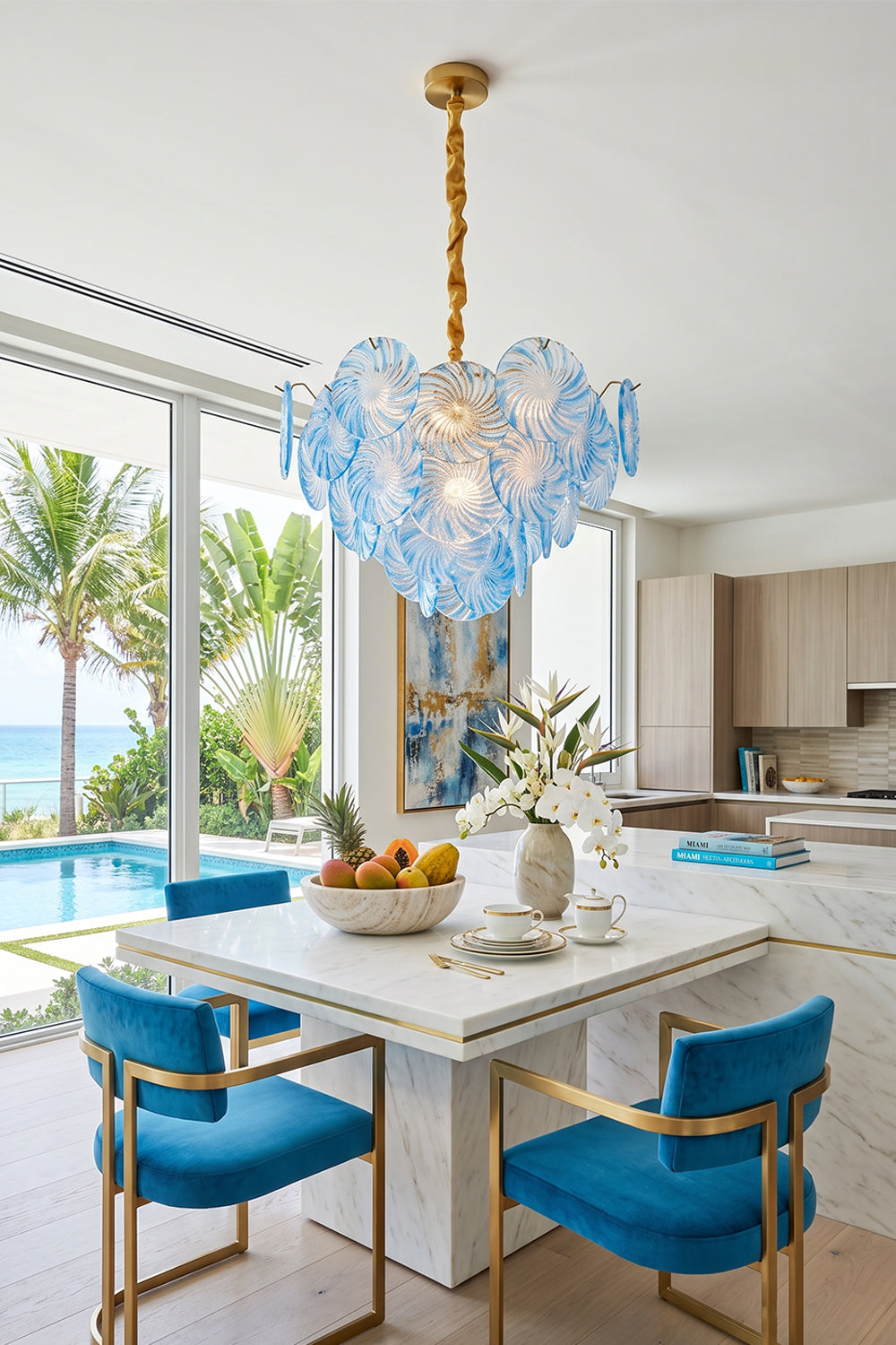 Amara Pink Glass Petal Chandelier | Dining Room Light | The sea-blue glass variant hanging elegantly over a modern white dining table paired with bright blue velvet chairs in a coastal home | Samu Lamp