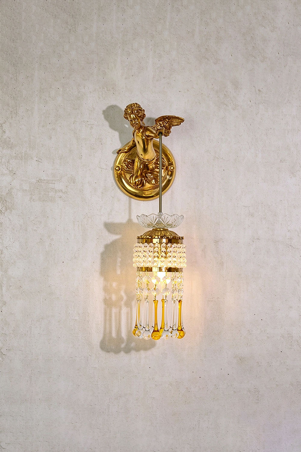 Ambrelise Cherub Wall Sconce | Brass and Crystal Glass | Front view mounted on grey plaster wall, illuminated | Samu Lamp