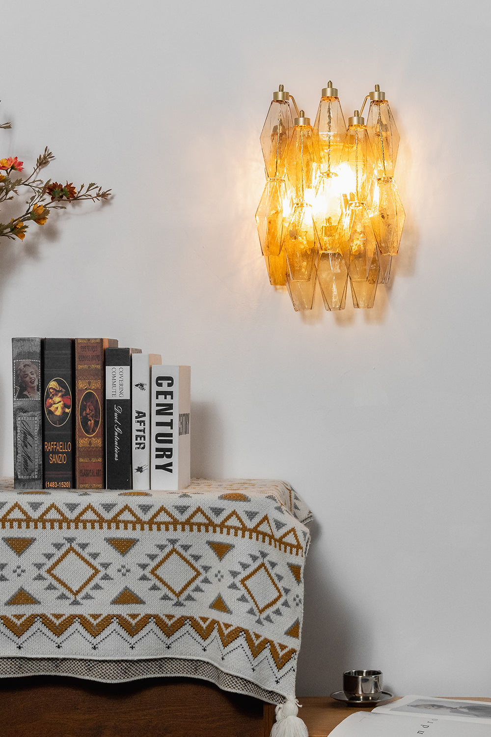 Lit amber polyhedral glass wall lamp | Metal, Glass | Mounted on a white wall above a console with books | Samu Lamp