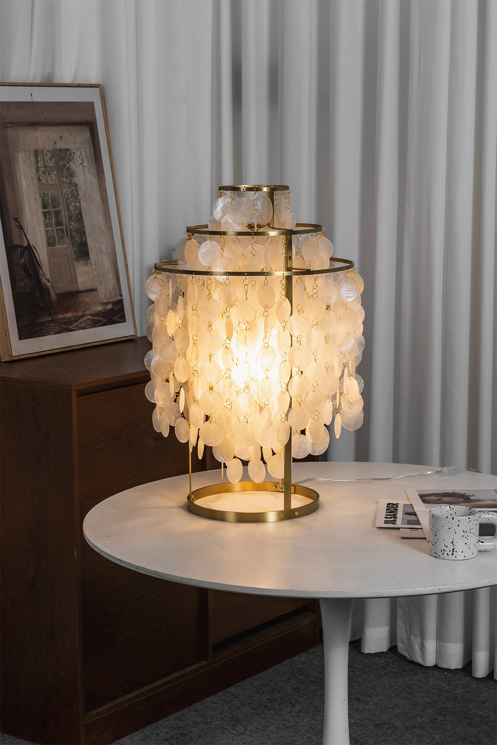 Aurelia Tiered Capiz Table Lamp | Capiz Shell, Metal | Lit on a modern white round table next to sheer white curtains in a minimalist room | Samu Lamp