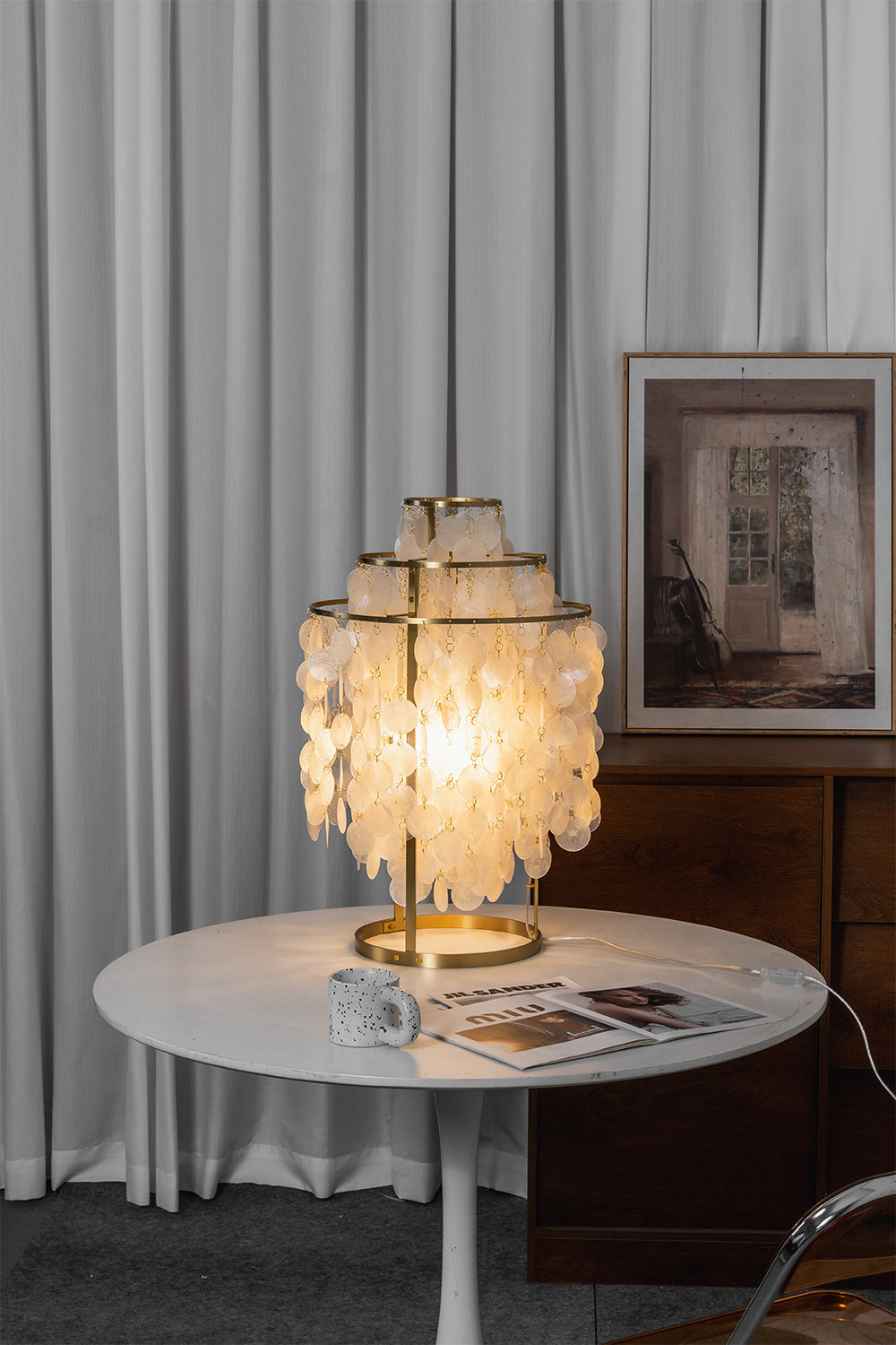 Aurelia Tiered Capiz Table Lamp | Capiz Shell, Metal | Vertical shot of the fully lit cascading capiz shell lamp on a white table against simple white curtains | Samu Lamp