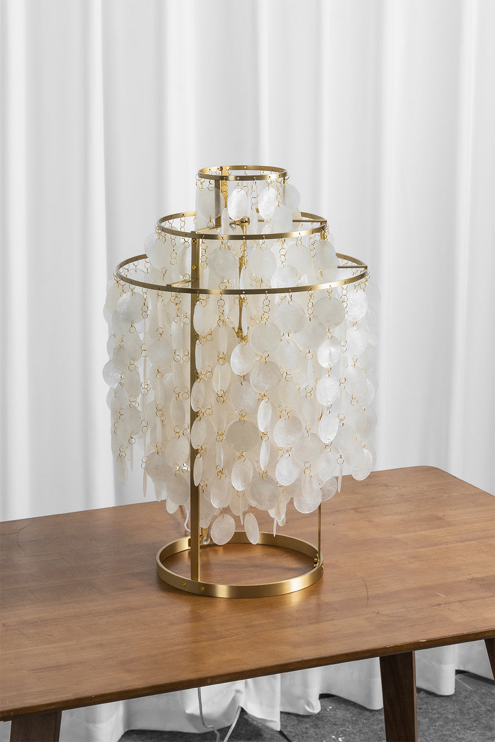 Aurelia Tiered Capiz Table Lamp | Capiz Shell, Metal | Staged unlit view of the tiered shell table lamp sitting on a rich wood surface next to sheer white blinds | Samu Lamp