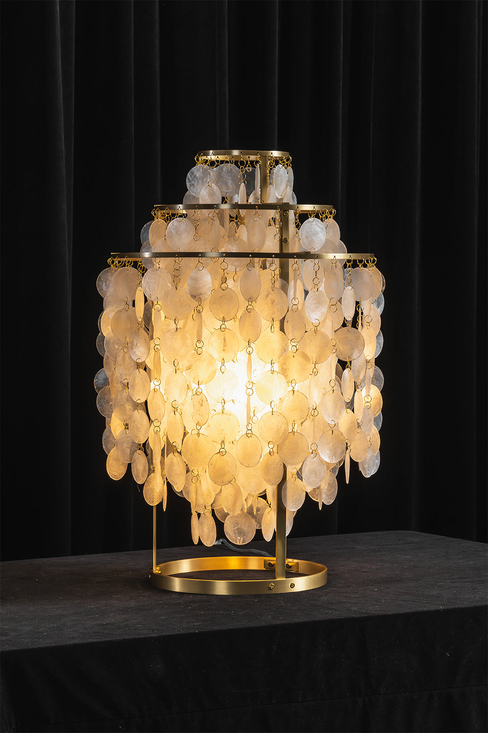 Aurelia Tiered Capiz Table Lamp | Capiz Shell, Metal | Dramatic dark studio shot of the fully illuminated tiered lamp radiating an intense golden glow against a black backdrop | Samu Lamp
