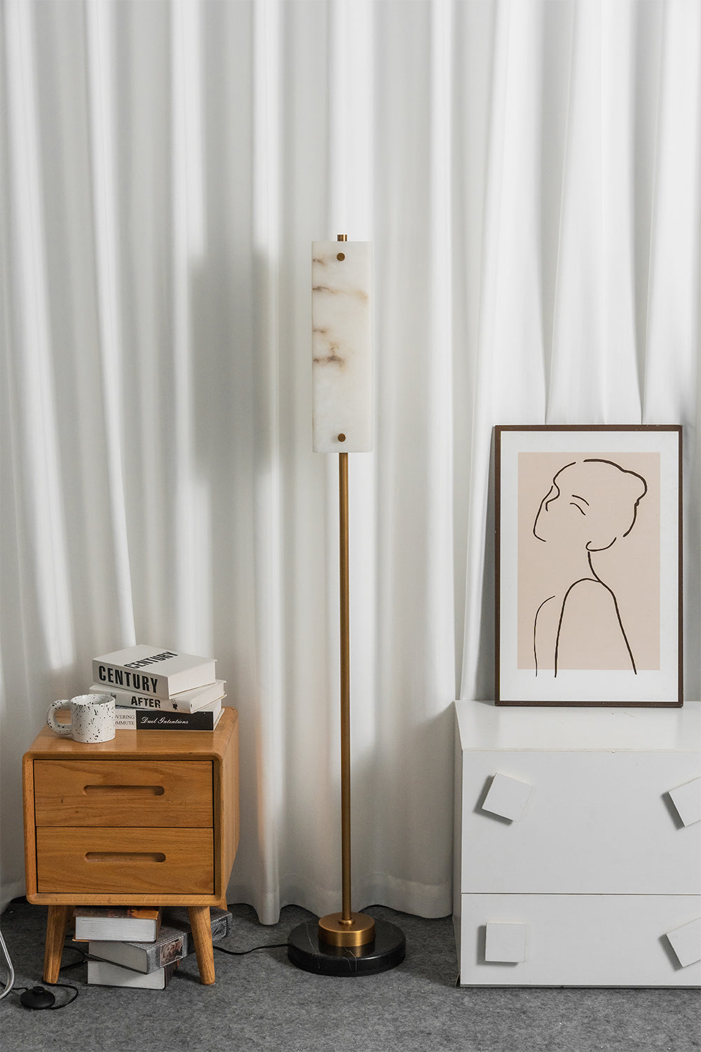 Aurelius Alabaster Brass Floor Lamp | Alabaster, Brass | Studio staged unlit view of the minimalist linear standing lamp set between a wooden nightstand and white drawers against a vertical pleated backdrop | Samu Lamp
