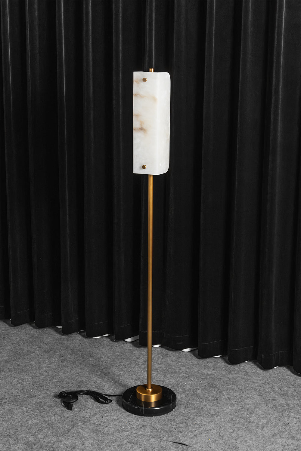 Aurelius Alabaster Brass Floor Lamp | Alabaster, Brass | Staged studio shot of the lit alabaster standing light radiating a warm yellow halo on a grey carpet against black vertical drapes | Samu Lamp