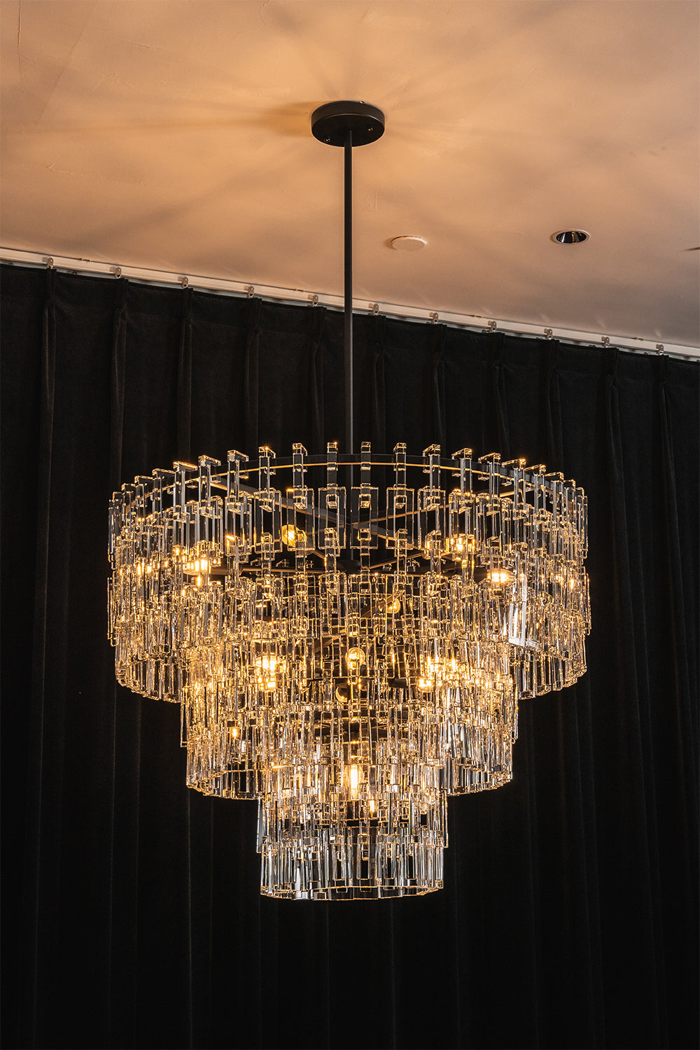Aven Tiered Glass Prism Chandelier | Glass, Metal | Full view of the illuminated tiered crystal fixture casting a soft golden glow against a dramatic dark wall backdrop | Samu Lamp