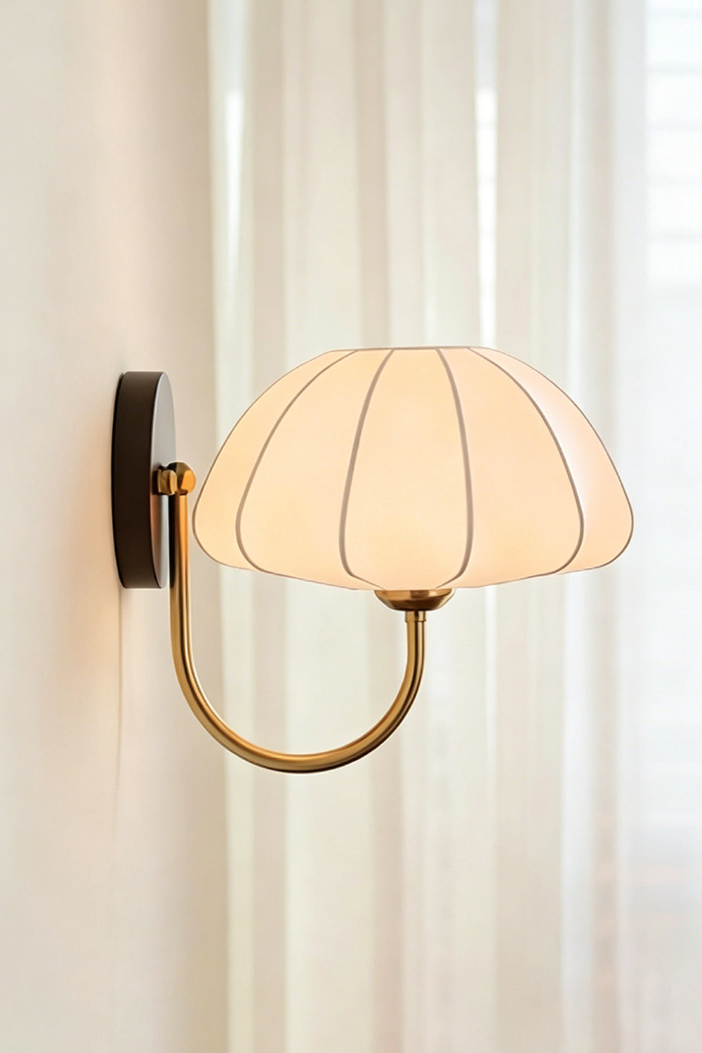 Baird Mid Century Pumpkin Wall Lamp | Metal, Fabric, Wood | Side profile view of the illuminated wall lamp next to soft white sheer curtains | Samu Lamp