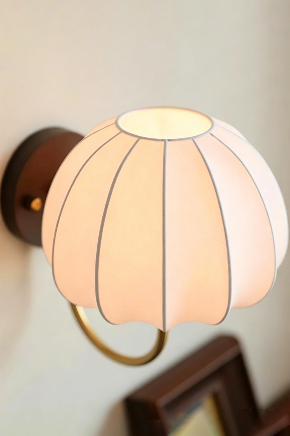 Baird Mid Century Pumpkin Wall Lamp | Metal, Fabric, Wood | High-angle shot of the fully lit pumpkin sconce mounted over a wooden picture frame | Samu Lamp
