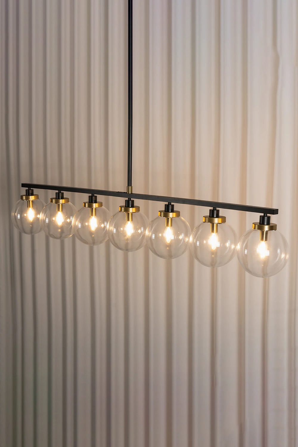 Brooks Linear Glass Globe Chandelier | Glass, Metal | Studio side angle view of the fully lit 7-globe linear suspension bar casting a bright warm light against a pleated white curtain background | Samu Lamp