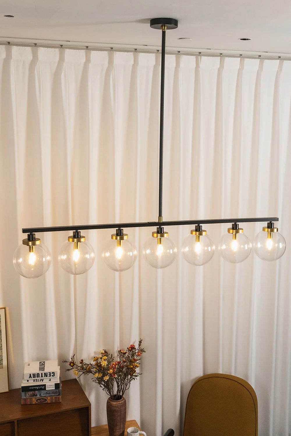 Brooks Linear Glass Globe Chandelier | Glass, Metal | Staged presentation of the fully illuminated multi-light linear island fixture glowing warmly against floor-to-ceiling white drapes | Samu Lamp