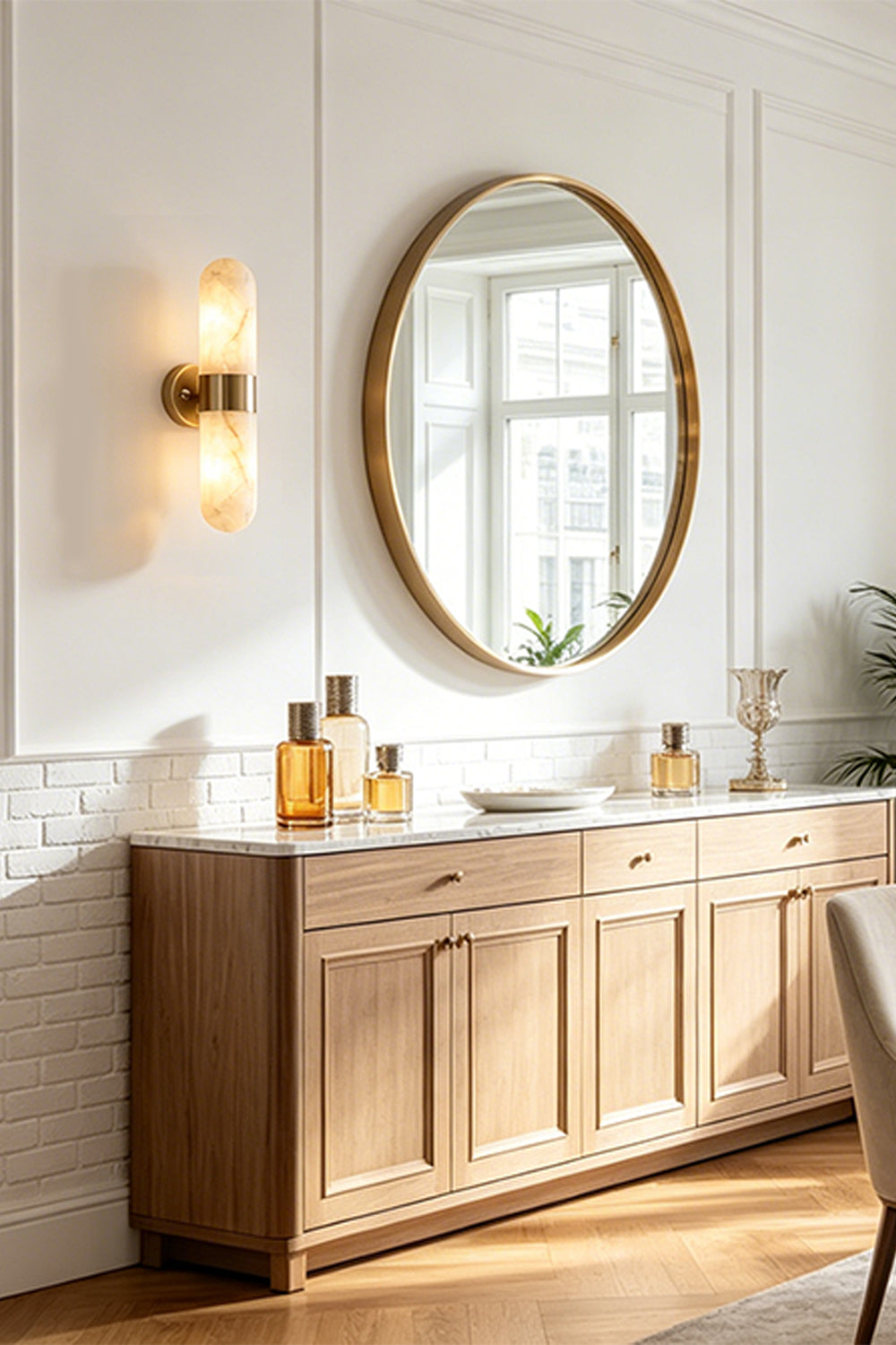 Calla Alabaster Capsule Wall Sconce | Alabaster, Brass | Mounted elegantly on a white wall next to a large round gold mirror and a wooden sideboard cabinet in a luxury hallway | Samu Lamp