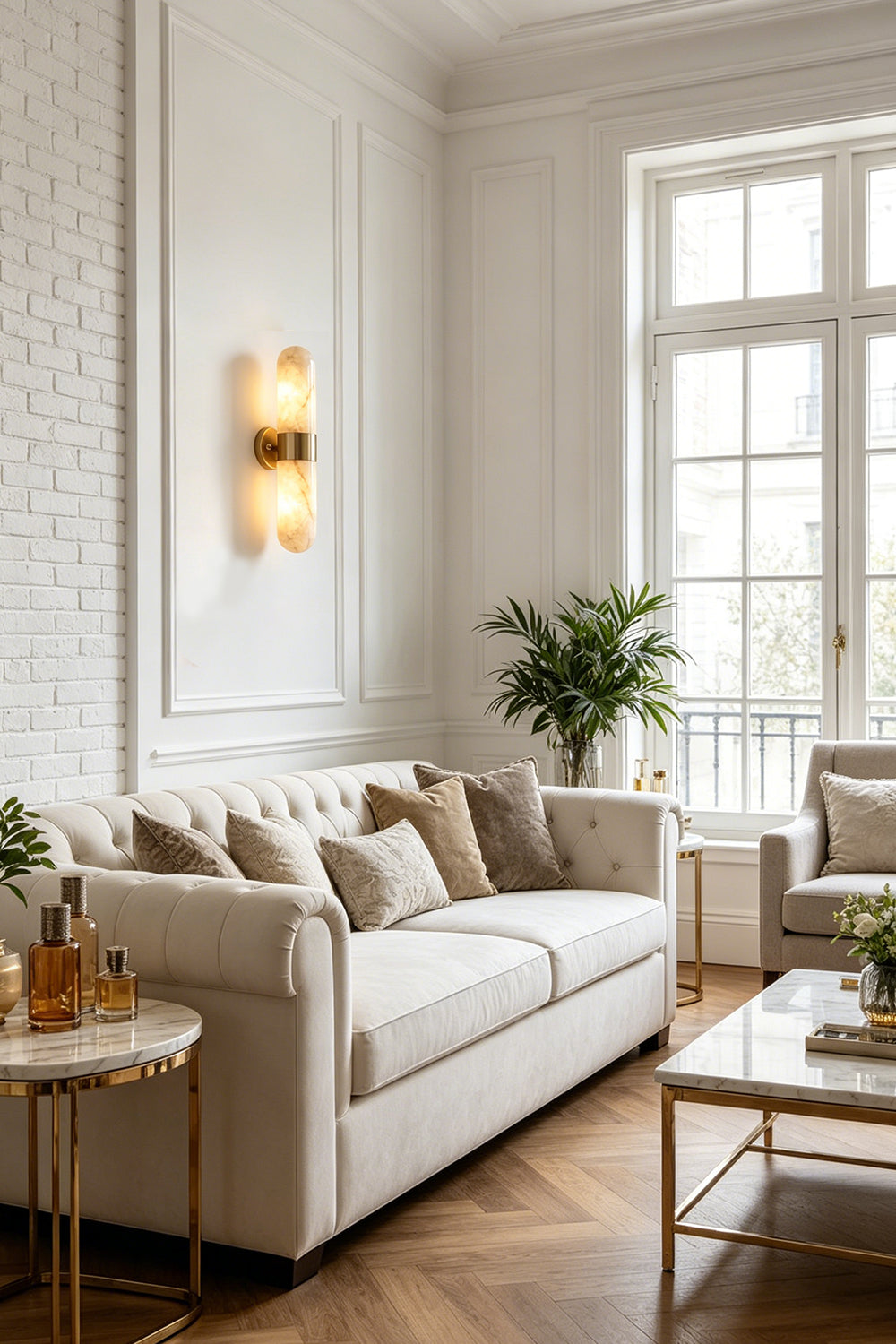 Calla Alabaster Capsule Wall Sconce | Alabaster, Brass | Providing a warm ambient glow on a white paneled wall in a sophisticated living room next to a cream fabric sofa | Samu Lamp