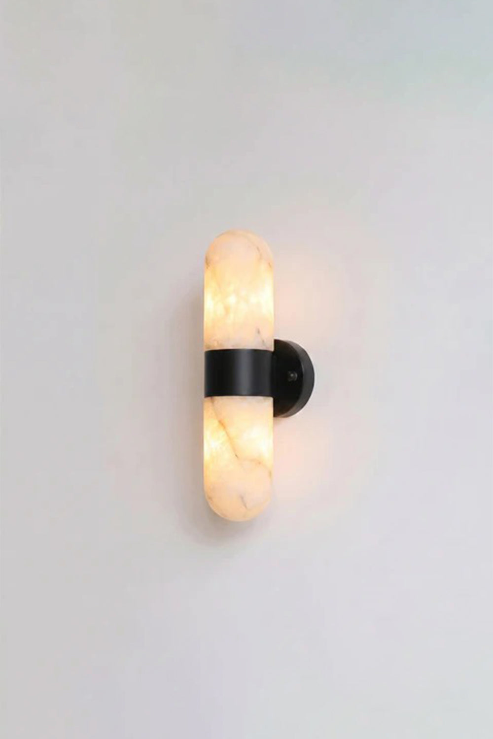 Calla Alabaster Capsule Wall Sconce | Alabaster, Metal | Studio front view of the fully lit matte black hardware variant casting a warm yellow halo against a plain background | Samu Lamp