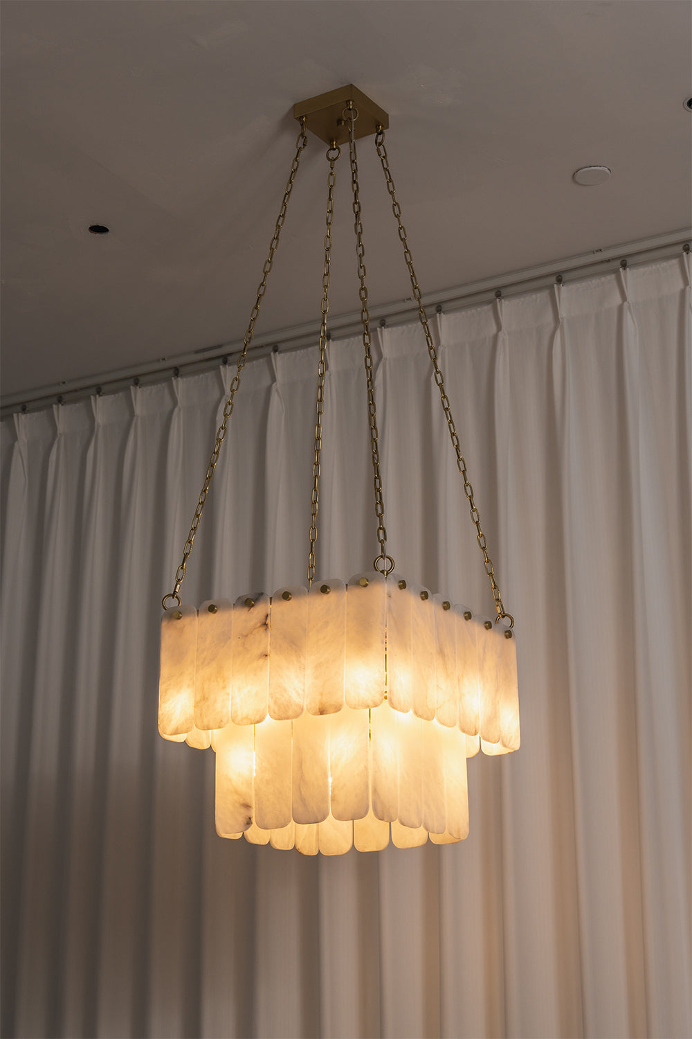 Callista Tiered Alabaster Chandelier | Alabaster, Metal | Studio centered view of the fully lit square tiered alabaster chandelier casting a beautiful warm architectural light against curtains | Samu Lamp
