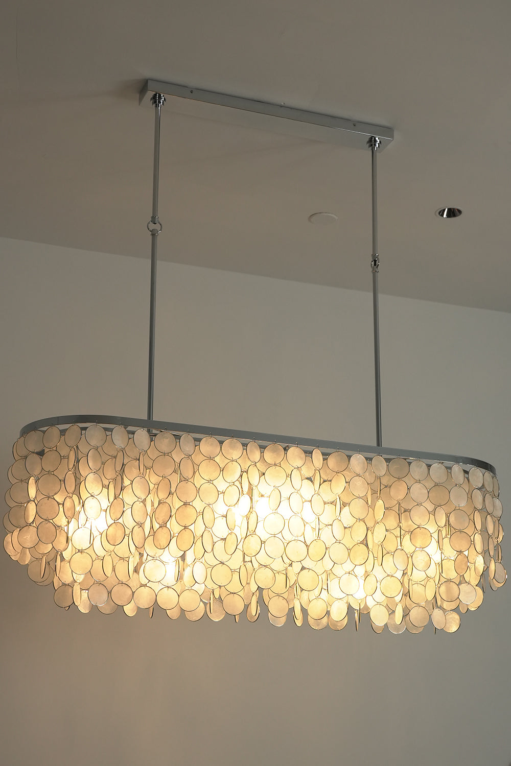 Lit Capiz shell elongated chandelier | Iron | Shell | Low angle view showing warm light filtering through shell discs | Samu Lamp