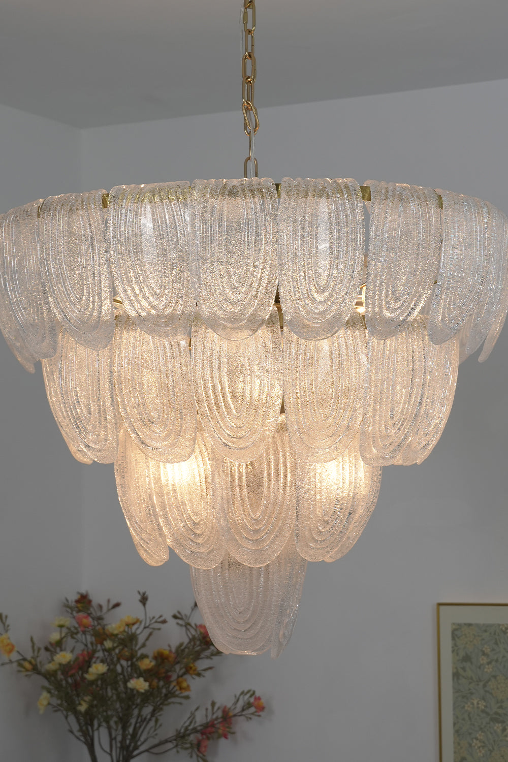 Lit cascading glass chandelier against neutral wall | Metal, Glass | Side perspective showing the tiered petal arrangement | Samu Lamp