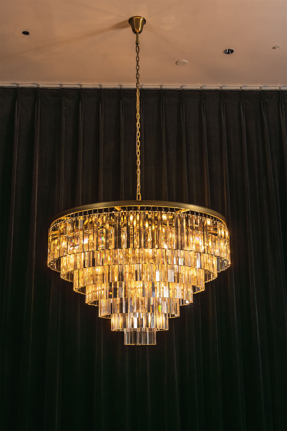 Cassian Tiered Prism Ring Chandelier | Clear Prism Glass and Brass | Full illuminated view displayed against a dark green curtain backdrop | Samu Lamp