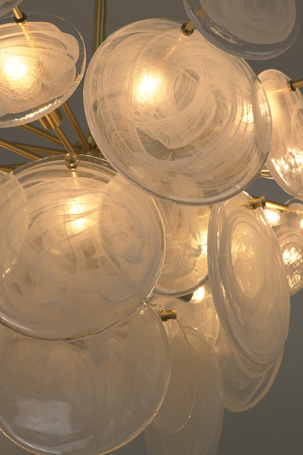 English: Celeste Glass Disc Chandelier | Glass, Metal | Macro scene view of overlapping lit textured glass discs emitting a cozy golden ambient light halo | Detail shot | Samu Lamp