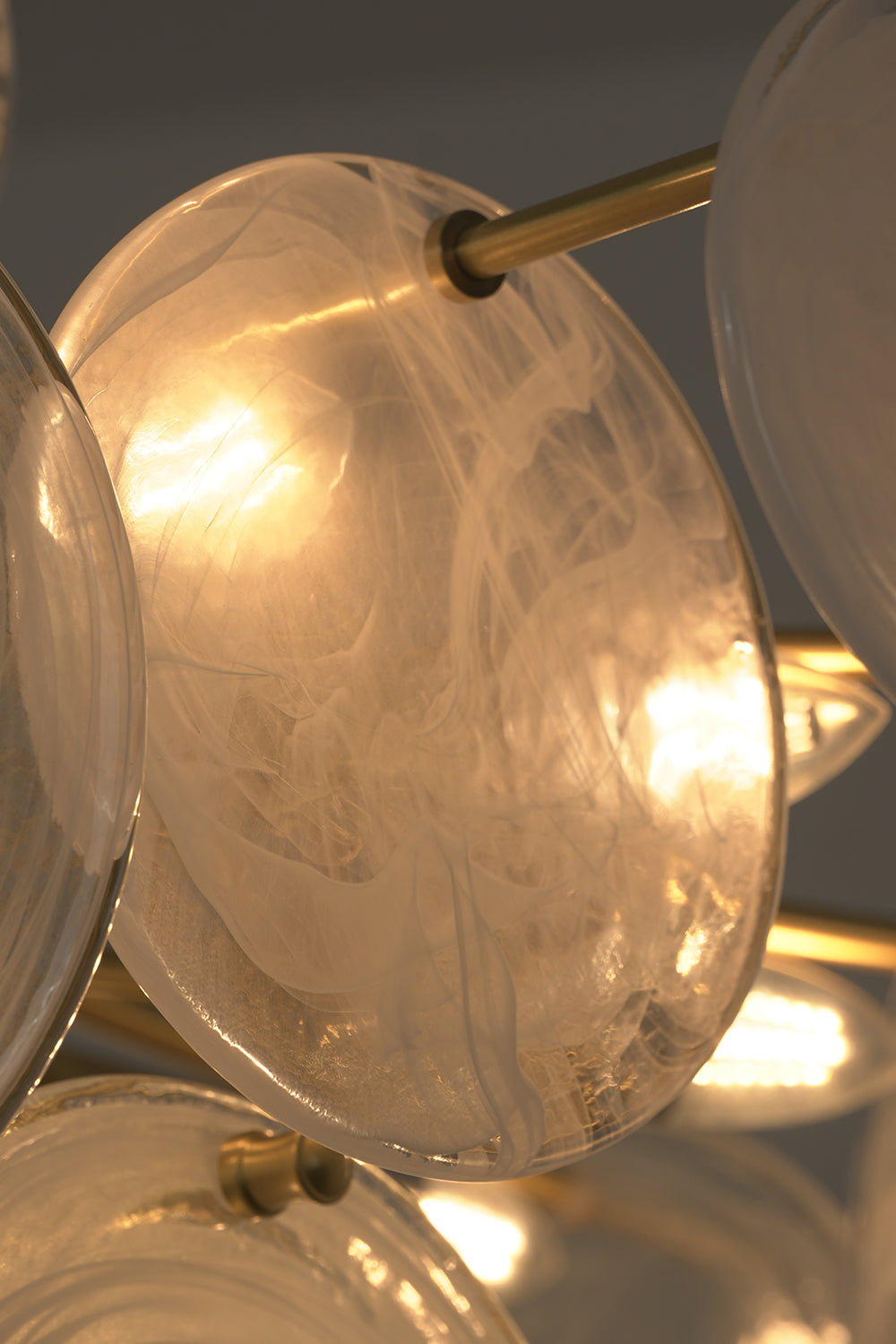 English: Celeste Glass Disc Chandelier | Glass, Metal | Close-up focus on an illuminated clear glass disc showing its organic ice-like surface pattern and warm light transmission | Detail shot | Samu Lamp