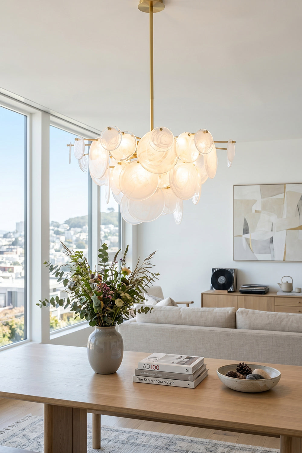 English: Celeste Glass Disc Chandelier | Glass, Metal | Positioned as an artistic centerpiece over a light oak wood table in a sunlit modern apartment lounge space | Samu Lamp