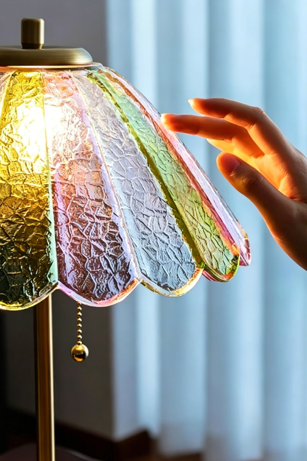 Clara Vintage Pastel Glass Table Lamp | Glass, Metal | A hand gently touching the scalloped edge of the fully illuminated colorful glass lampshade near a window | Samu Lamp