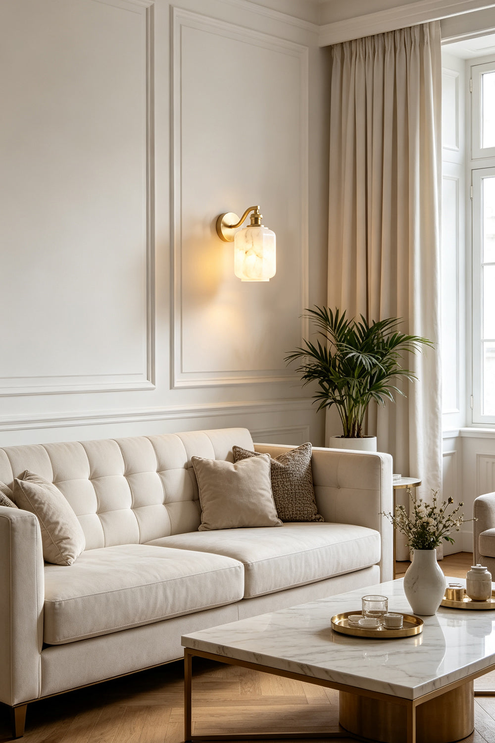 Cordelia Scalloped Alabaster Wall Sconce | Natural Alabaster and Brushed Brass | Mounted on a white wainscoting wall casting a warm glow behind a classic cream fabric sofa | Samu Lamp