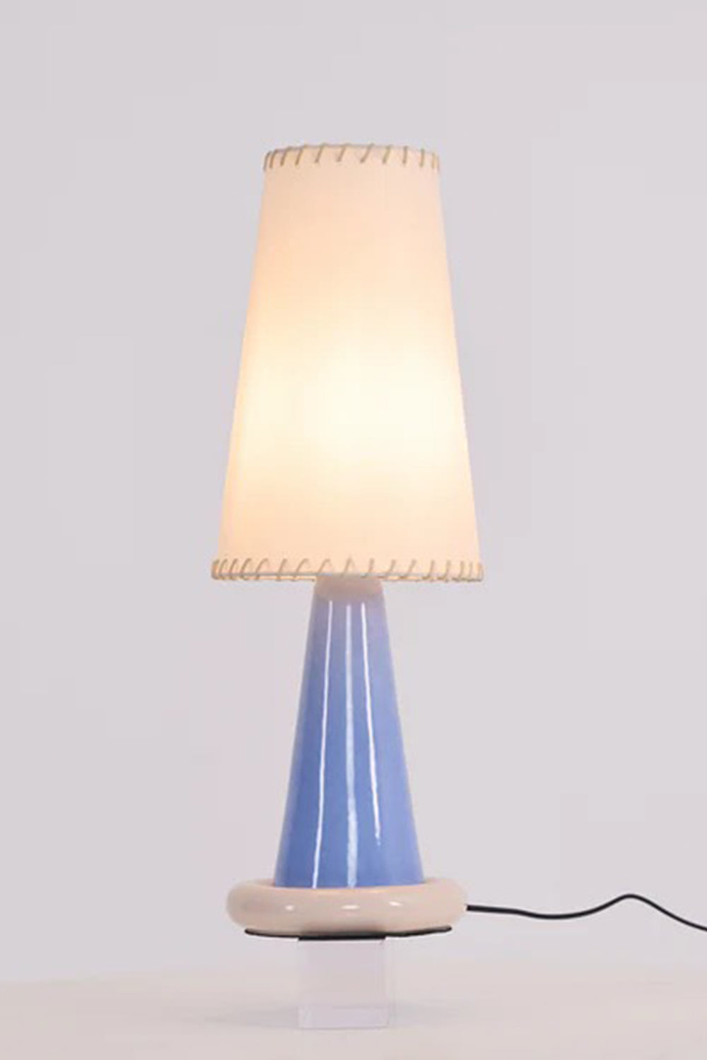 Dolce Ribbon Ceramic Table Lamp | Ceramic, Fabric | Studio view of the fully illuminated tall white shade variant casting a soft warm white glow against a plain background | Samu Lamp