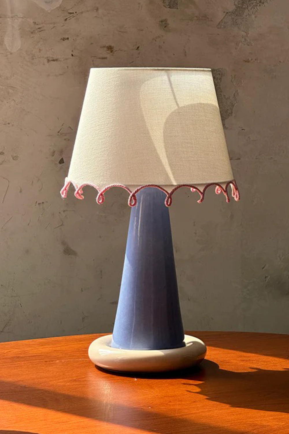Dolce Ribbon Ceramic Table Lamp | Ceramic, Fabric | Lit product resting on a wooden surface under warm dramatic window sunlight stripes against a textured wall | Samu Lamp