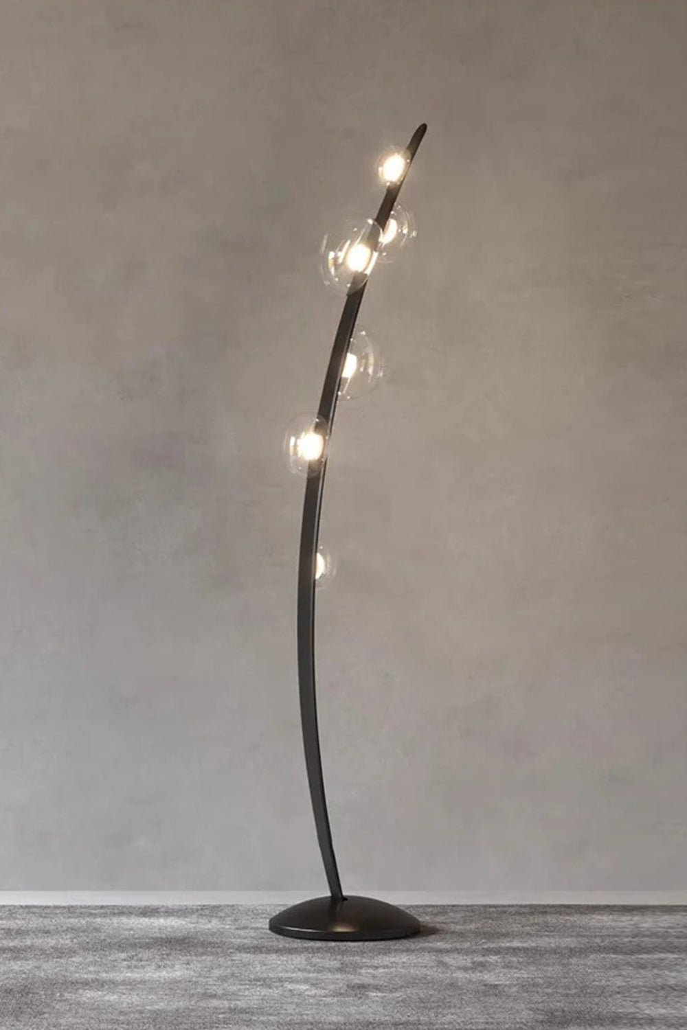 Drosia Dewdrop Floor Lamp | Glass, Metal | Studio view of the fully illuminated minimalist black floor lamp radiating a cozy golden light alignment against a plain grey textured wall | Samu Lamp
