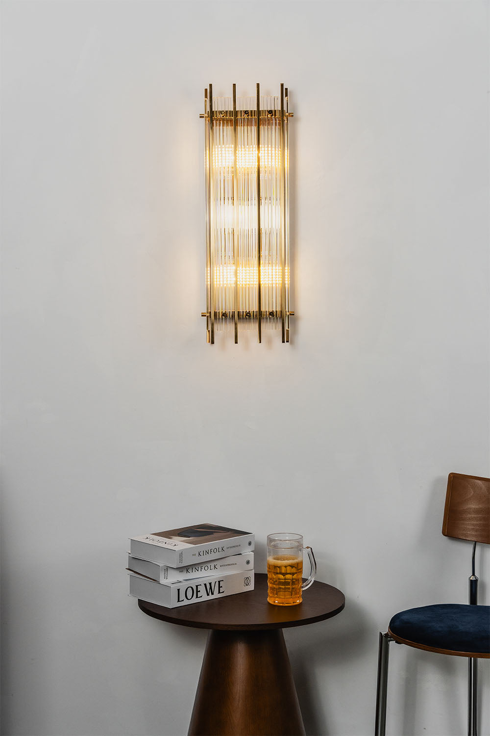 Drystan Ribbed Glass Brass Wall Lamp illuminating a wall above a side table with books and a drink.