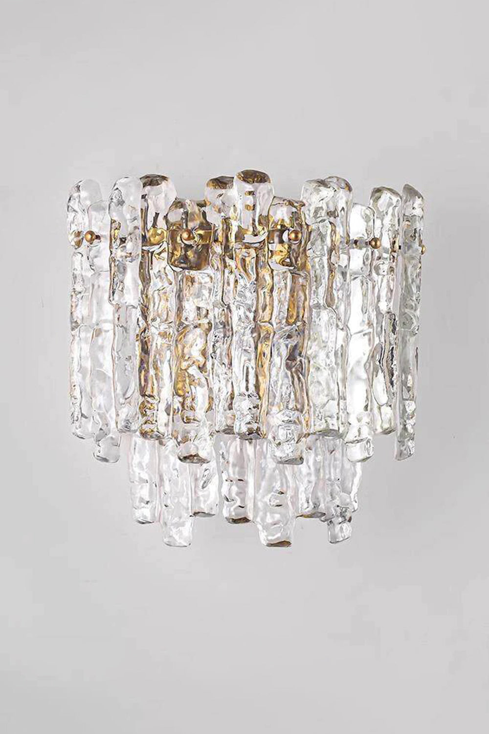 Elara Textured Cast Glass Wall Lamp | Textured Cast Glass, Polished Brass | Medium angle shot of the illuminated lamp against a neutral grey wall, demonstrating its shape and the dramatic textured glass effect in a corridor or entryway | Samu Lamp