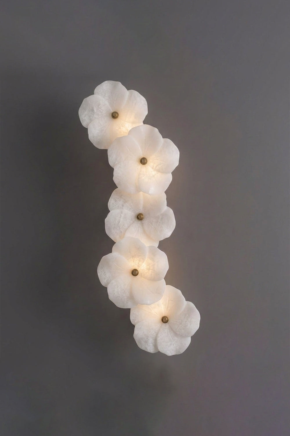 Elowen Alabaster Blossom Wall Light | Studio Lit View | Studio front shot of the fully illuminated vertical floral stone sconce glowing warmly against a solid dark grey backdrop | Samu Lamp