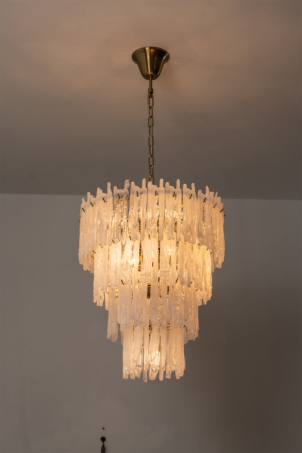 Ethereal Icicle Waterfall Chandelier | Glass, Metal | Full view of the fully illuminated tiered chandelier casting a dramatic warm light against a plain wall | Samu Lamp