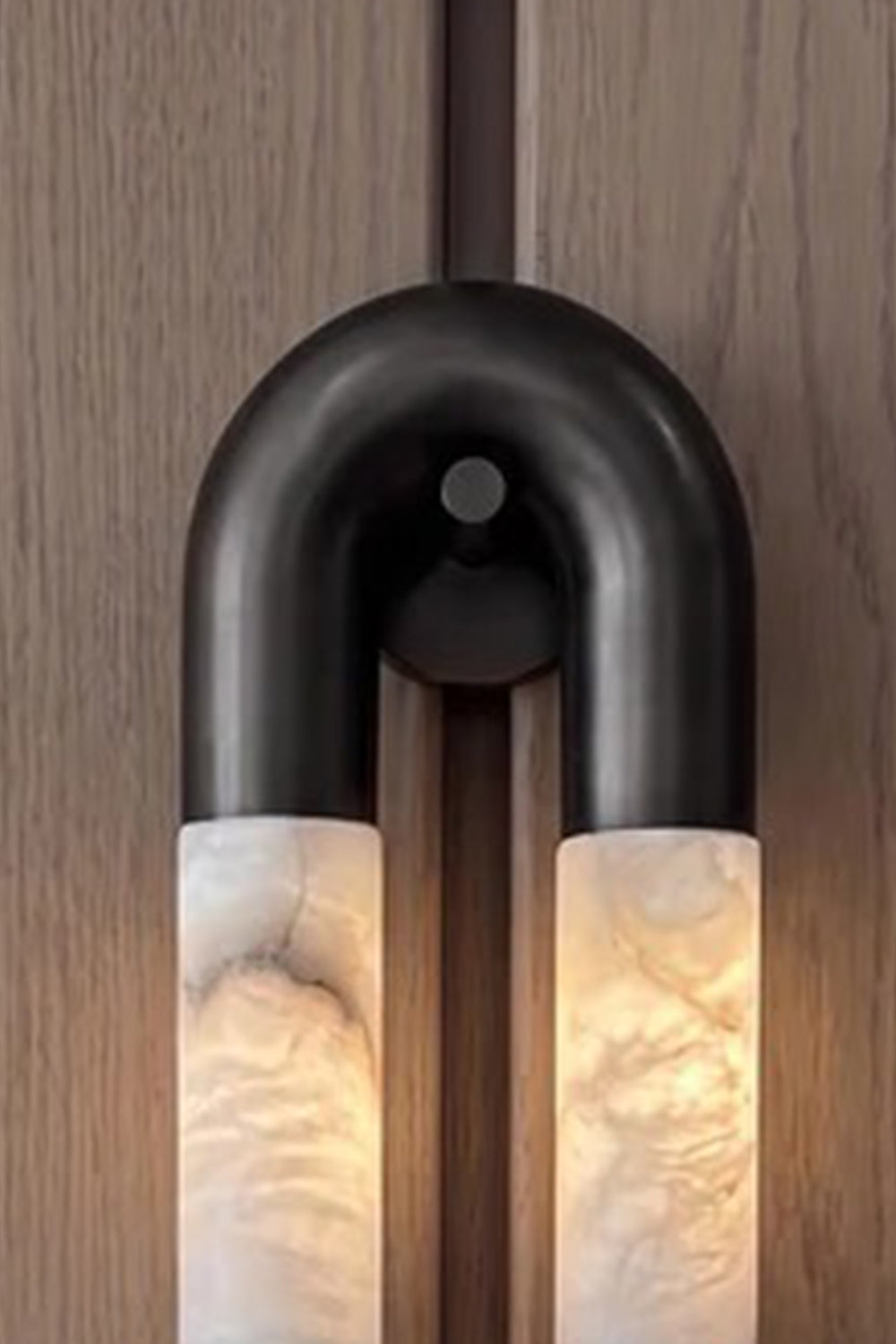 Evadne Alabaster Link Wall Sconce | Alabaster, Metal | Close-up focus on the top curved matte black metallic arch connecting seamlessly to the glowing natural veined alabaster cylinders | Detail shot | Samu Lamp
