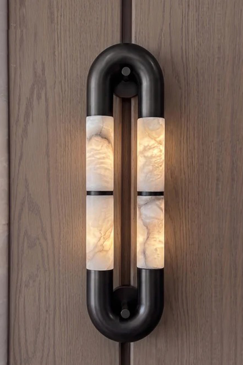 Evadne Alabaster Link Wall Sconce | Alabaster, Metal | Sconce fully lit up on a natural dark wood grain panel background, showcasing the beautiful play of light and shadow | Samu Lamp