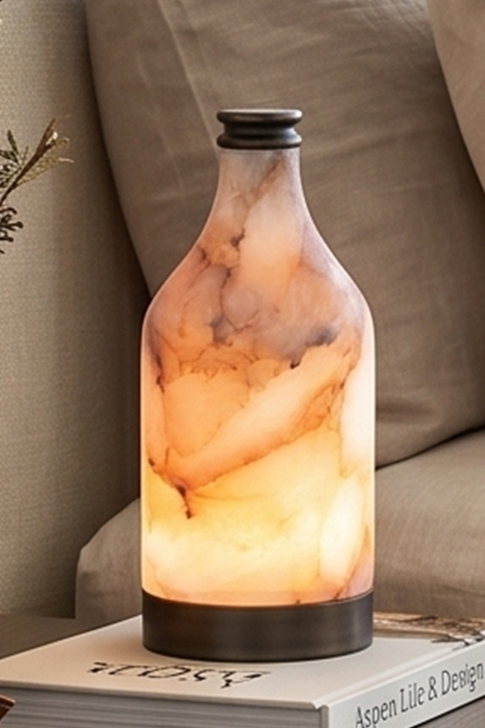 Evander Alabaster Bottle Table Lamp | Alabaster, Metal | Detailed staged view of the illuminated natural alabaster lamp resting on a book next to a cozy fabric lounge couch | Samu Lamp