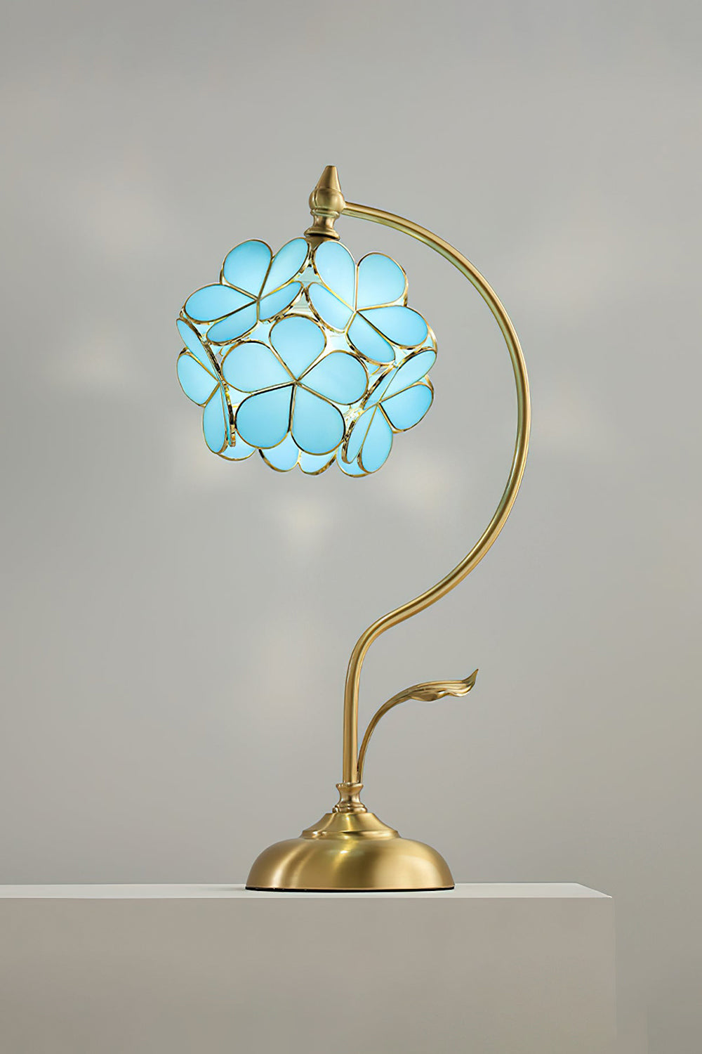 Evanthe Blossom Stained Glass Table Lamp | Stained Glass, Metal | Studio front view of the vibrant sky blue variant fully illuminated on a white pedestal block against a grey wall | Samu Lamp
