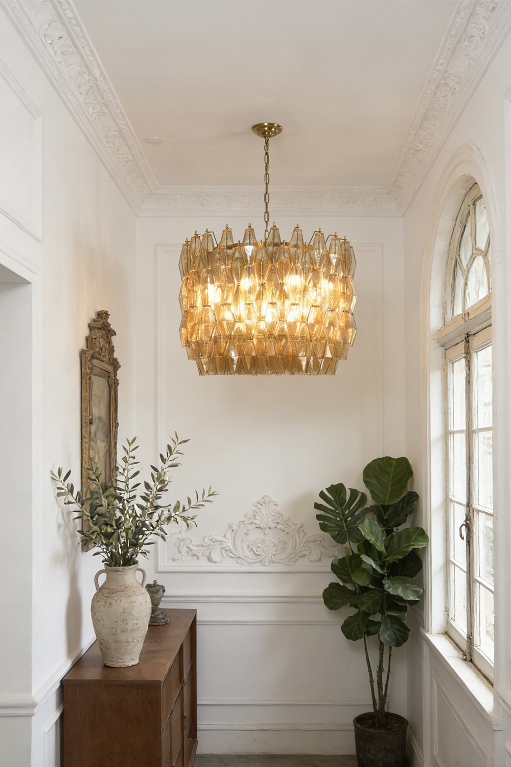 Luxurious amber glass drum chandelier | Metal, Glass | Hanging in a hallway with white wainscoting and plants | Samu Lamp