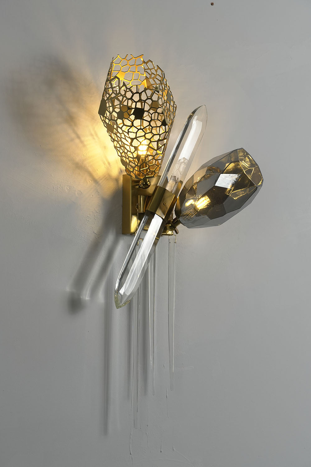 Faucenne Pierced Shade Crystal Spear Wall Lamp | Cut Crystal and Brass | Front view glowing with warm light against a grey wall backdrop | Samu Lamp