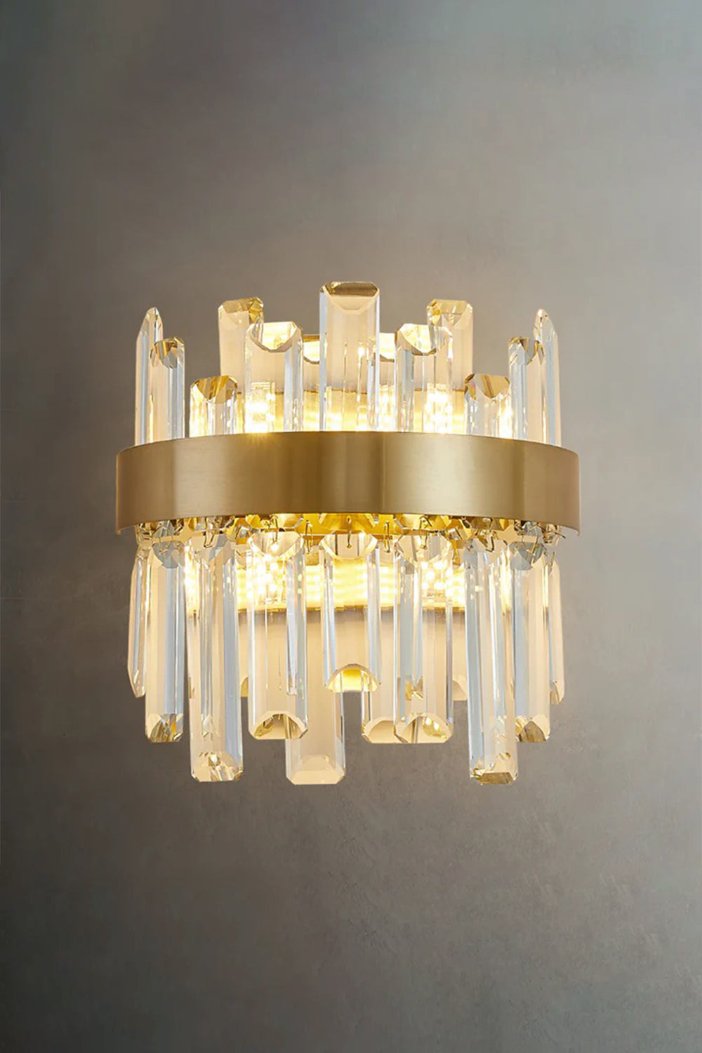 Glacia Tiered Crystal Statement Wall Lamp | Crystal, Metal | Studio front view of the fully illuminated crystal sconce casting a rich warm light halo on a dark textured grey background wall | Samu Lamp