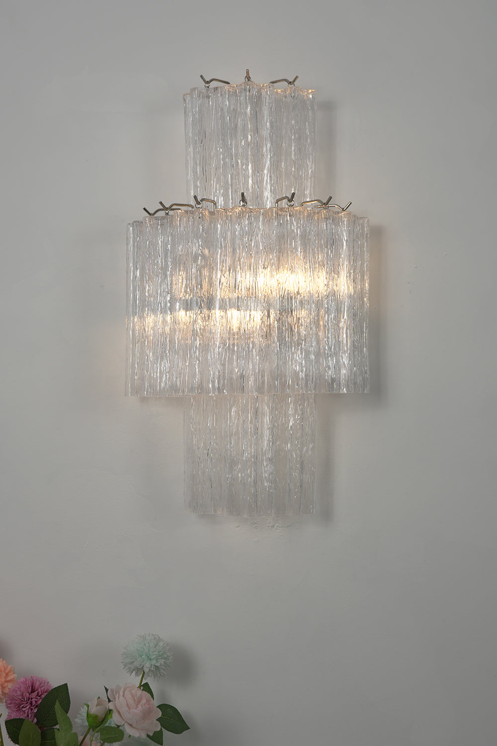 Lit glacier glass wall light over a vase of flowers | Metal, Glass | Close-up view against a grey wall | Samu Lamp