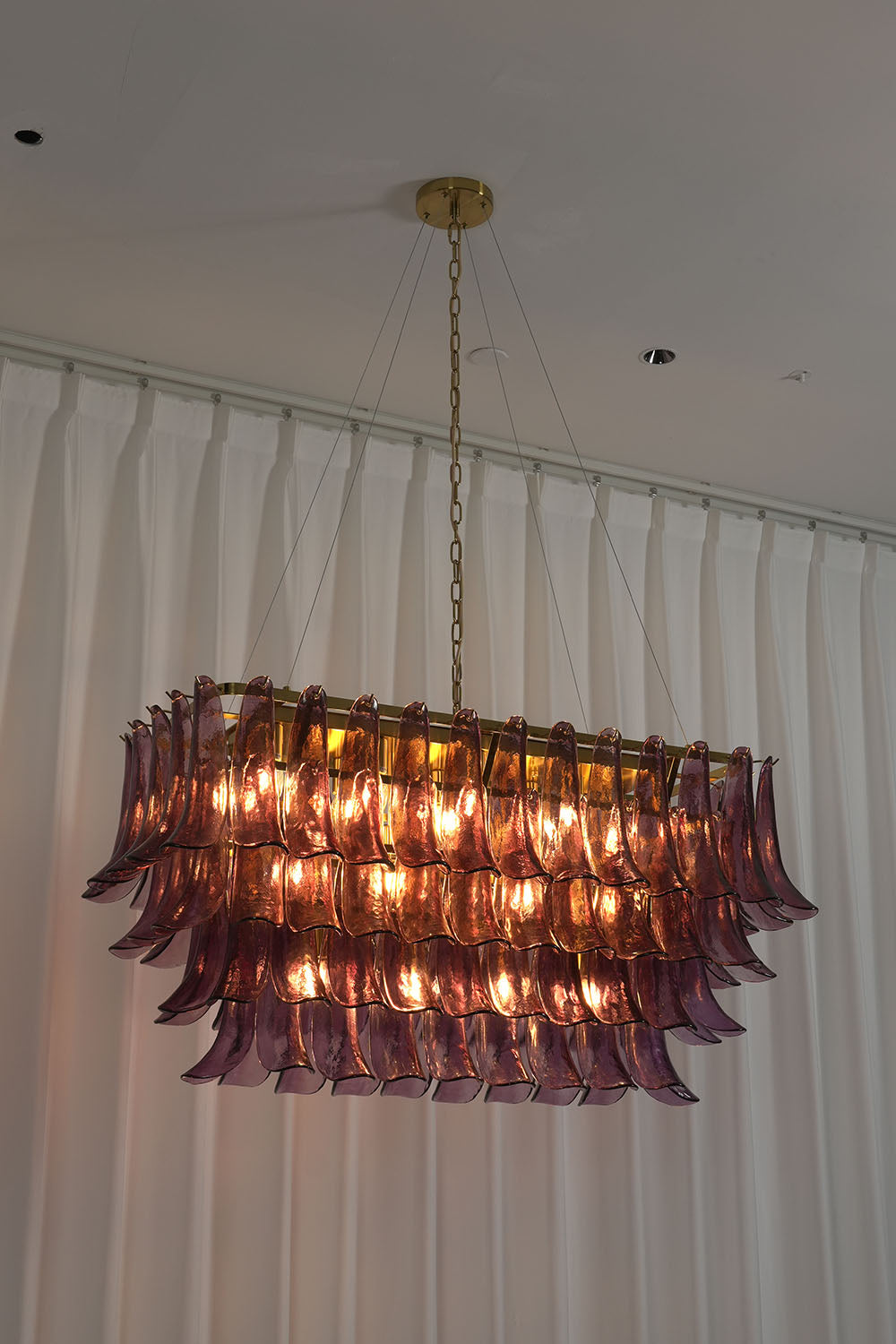 Ilvesse Amethyst Glass Petal Rectangular Chandelier | Amethyst Glass and Brass | Real-life view glowing warmly against white pleated window drapes | Samu Lamp