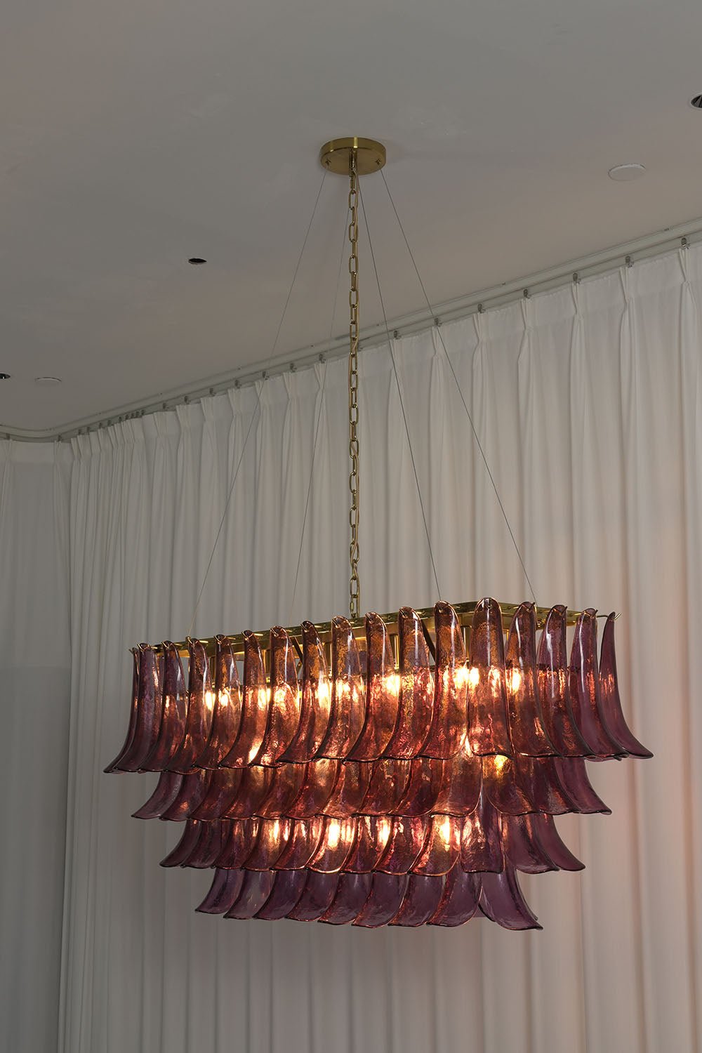 Ilvesse Amethyst Glass Petal Rectangular Chandelier | Amethyst Glass and Brass | Real-life installation view glowing softly against a plain grey wall | Samu Lamp