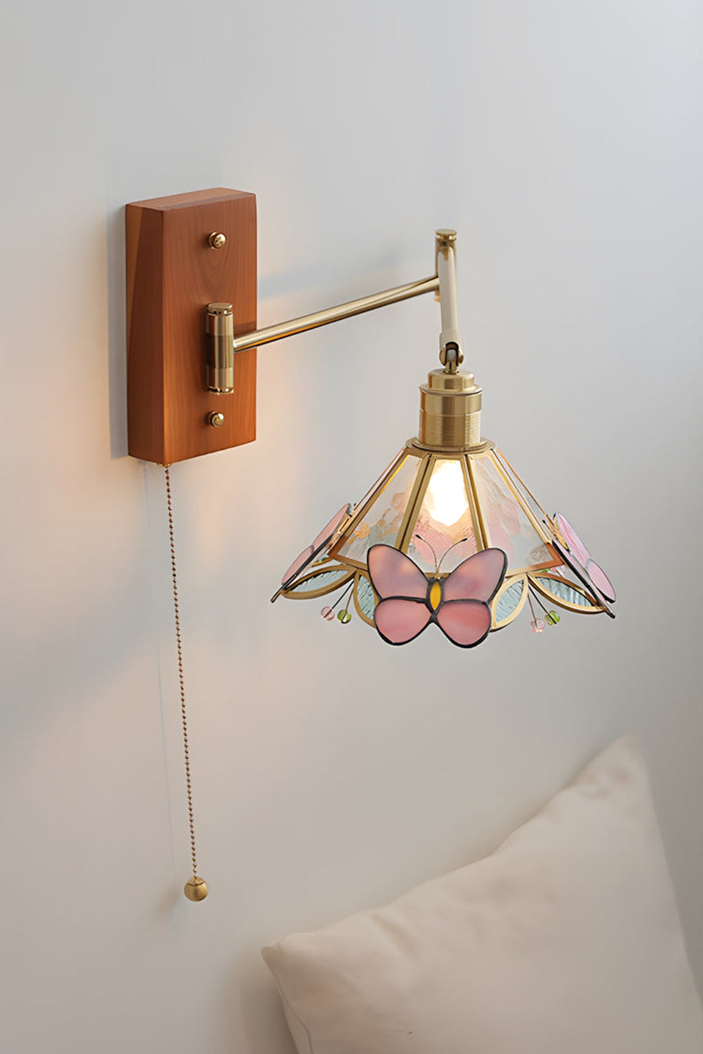 Iris Butterfly Stained Glass Wall Lamp | Stained Glass, Metal, Wood | Mounted on a plain white wall above a soft cream pillow, showcasing the full extension of the gold brass swing arm and the hanging pull chain | Samu Lamp
