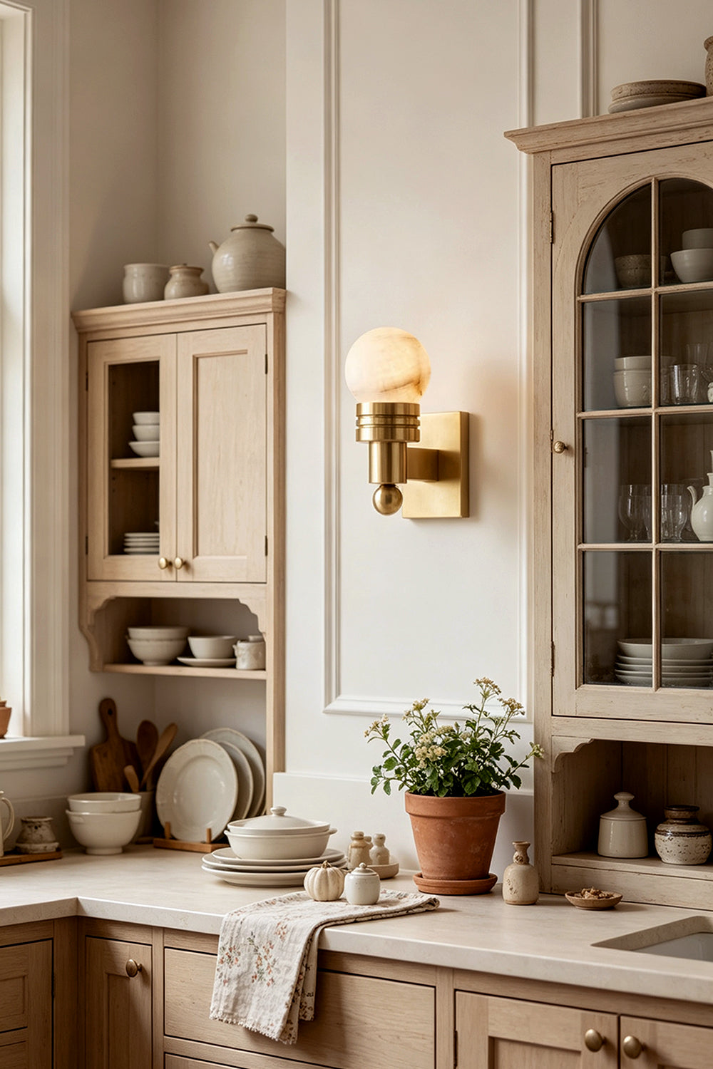 Isolde Alabaster Orb Wall Sconce | Natural Alabaster and Aged Brass | Fixed on the wall of a charming cottage kitchen above a counter with a small plant | Samu Lamp