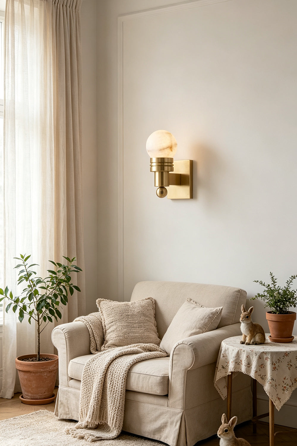 Isolde Alabaster Orb Wall Sconce | Natural Alabaster and Aged Brass | Styled on a living room wall above a comfortable beige fabric sofa and a potted plant | Samu Lamp