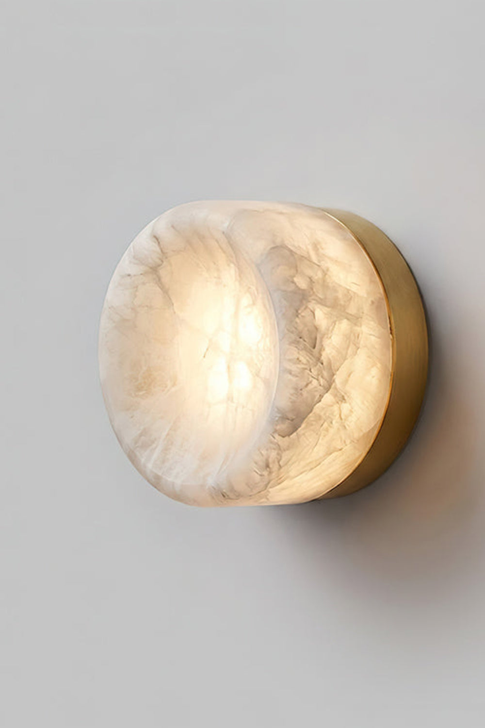 Isolde Concave Alabaster Disc Wall Sconce | Natural Alabaster and Black Metal | Studio close-up shot of the lit sconce featuring a matte black finish base on a light wall | Samu Lamp
